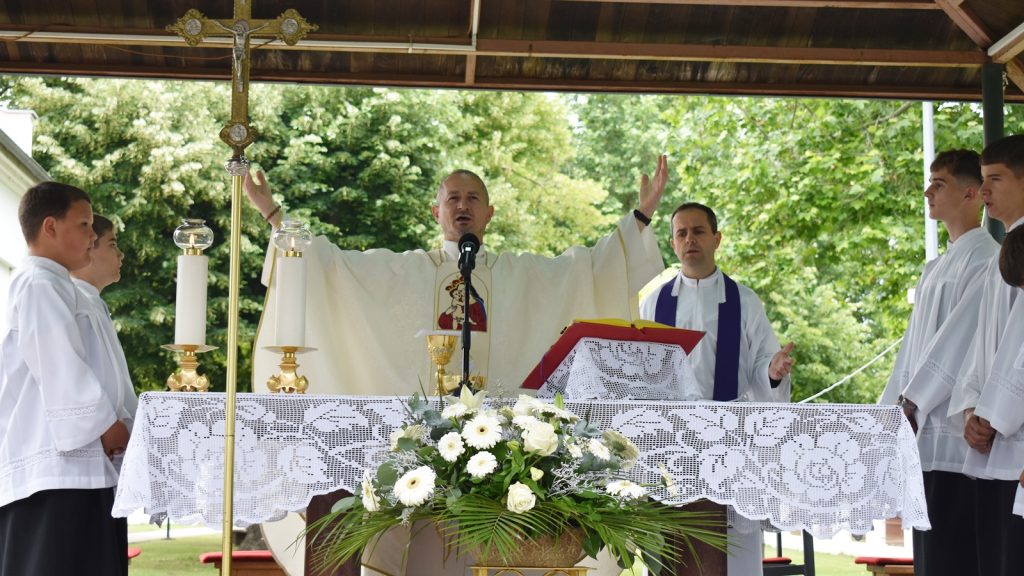 Priest raises arms at an outdoor altar with white lace cloth, flanked by altar servers and candles during Mass