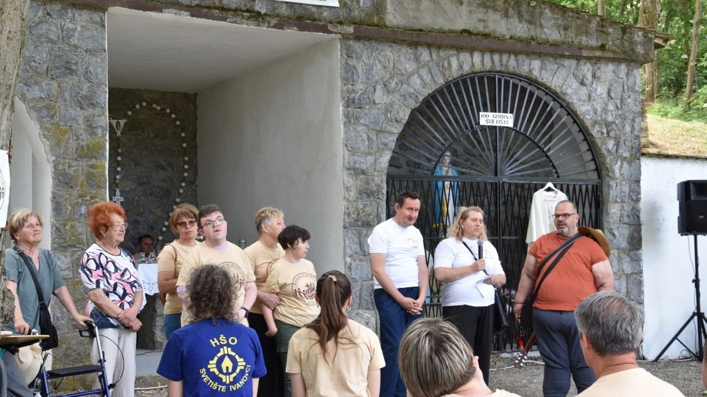 Group of people standing outdoors near a stone arch, one man speaks into a microphone while others listen.