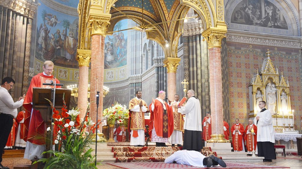 Catholic ceremony in a lavish church with priests in red vestments at an ornate altar; a person lies on the carpet in front of them.
