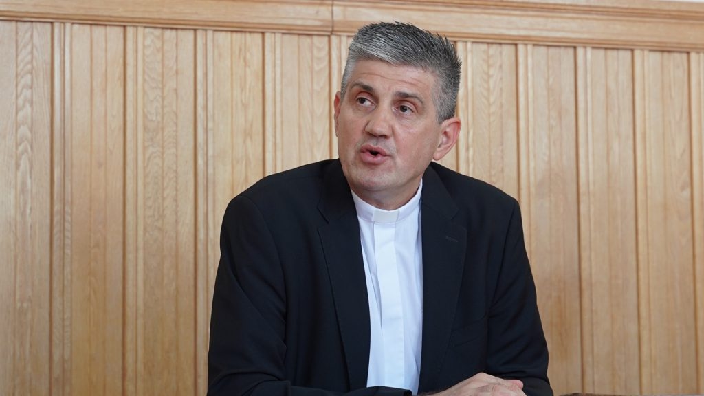 Man in a black suit with a clerical collar seated at a table, speaking in front of a wood-paneled wall.