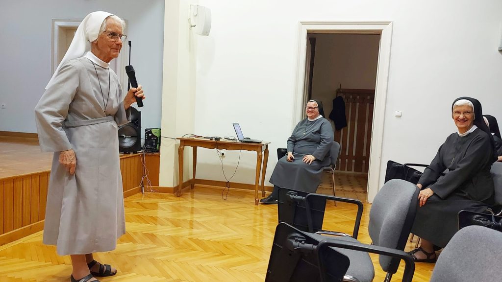 Older nun in a gray habit speaks into a handheld microphone on a small stage to seated nuns in the room.
