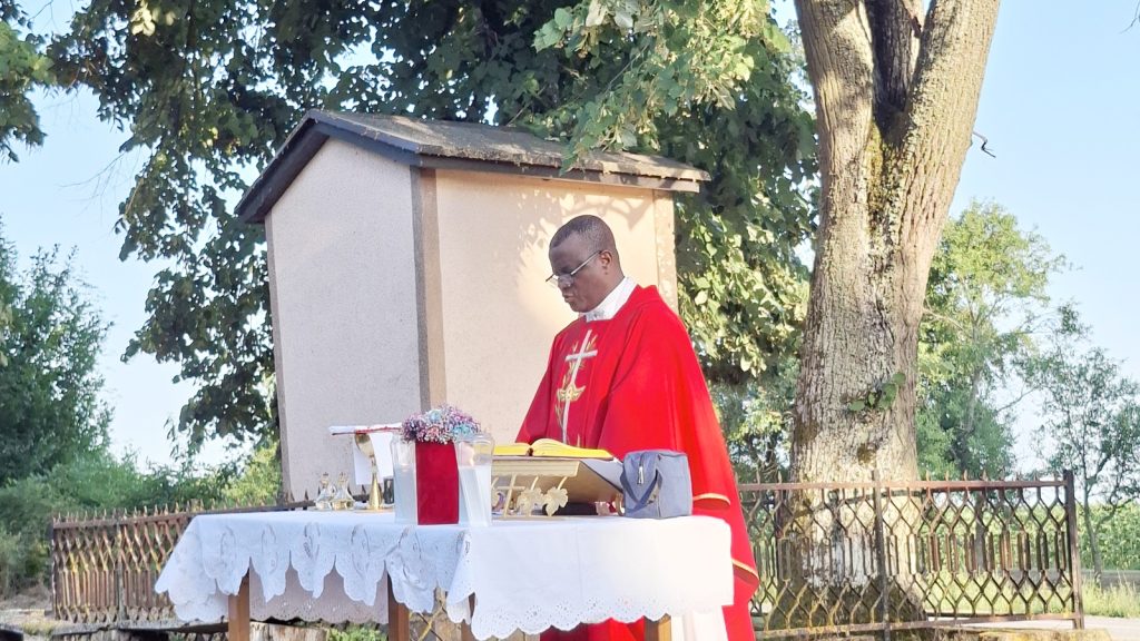 Priest in red vestments conducts an outdoor Mass at a table-alter with a Bible, chalice, and flowers under a tree.