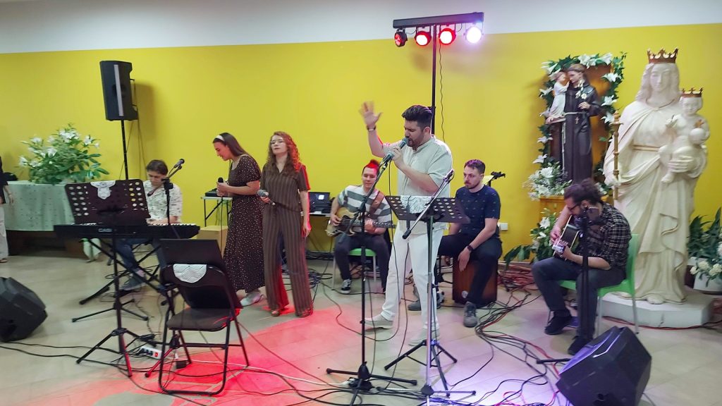 A music group performs indoors on a yellow wall stage with multiple singers and instrumentalists including a keyboardist, guitarist, and a player on percussion.