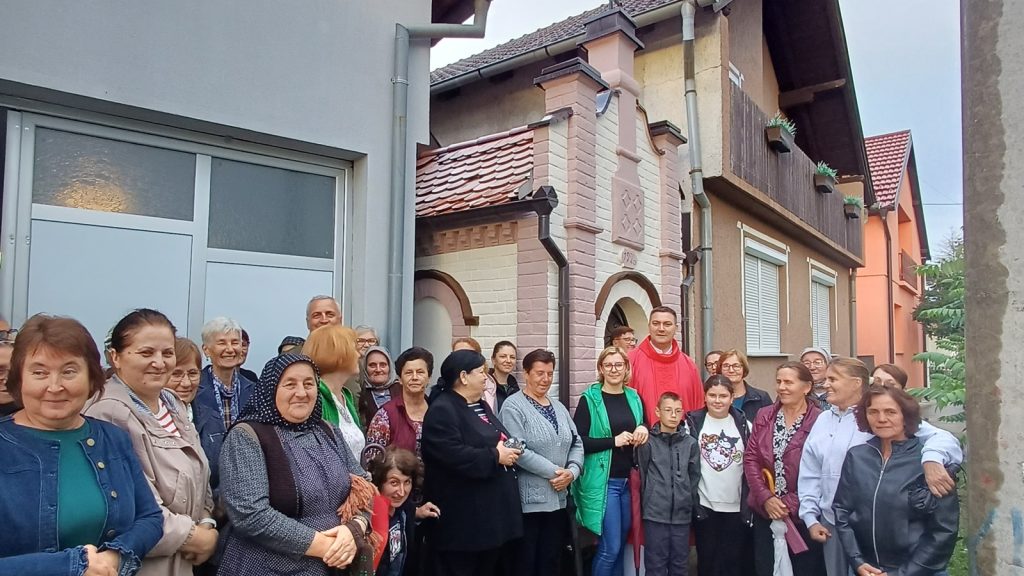 Large group of people posing together outside a row of colorful houses, smiling at the camera in a community gathering.