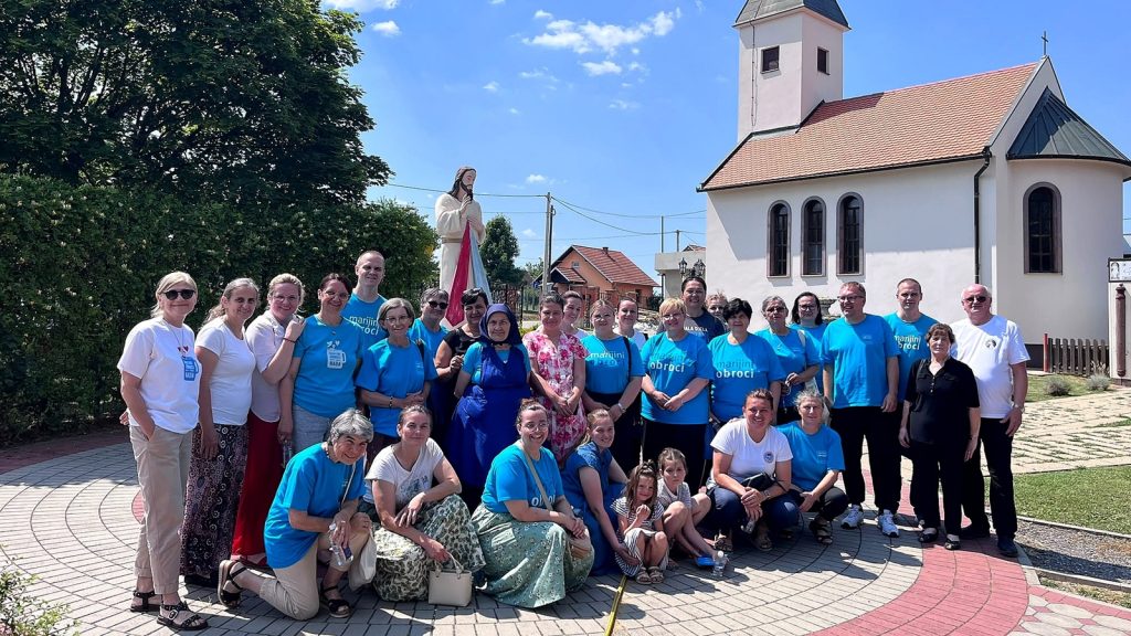 Large group of people posing for a photo outside a church on a sunny day, many wearing matching blue t-shirts.
