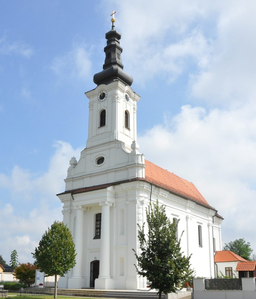White church with a tall, dark multi-tiered steeple topped by a gold cross against a bright blue sky, clock faces on the tower faces visible from below.