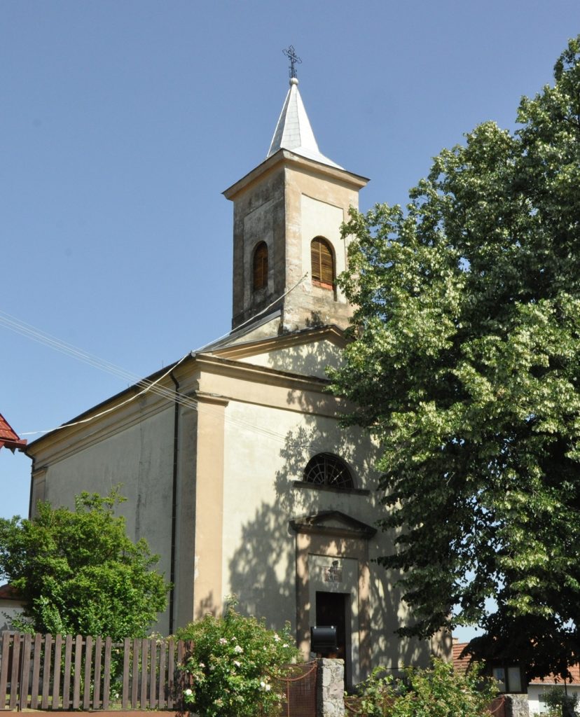 Church with tall steeple and cross against a clear blue sky, partially framed by a leafy tree in the foreground.