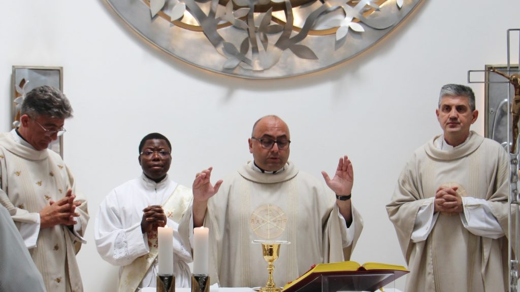 Priest with raised hands blessing at the altar during Mass, with candles, chalice, and open Bible nearby, flanked by fellow clergy.