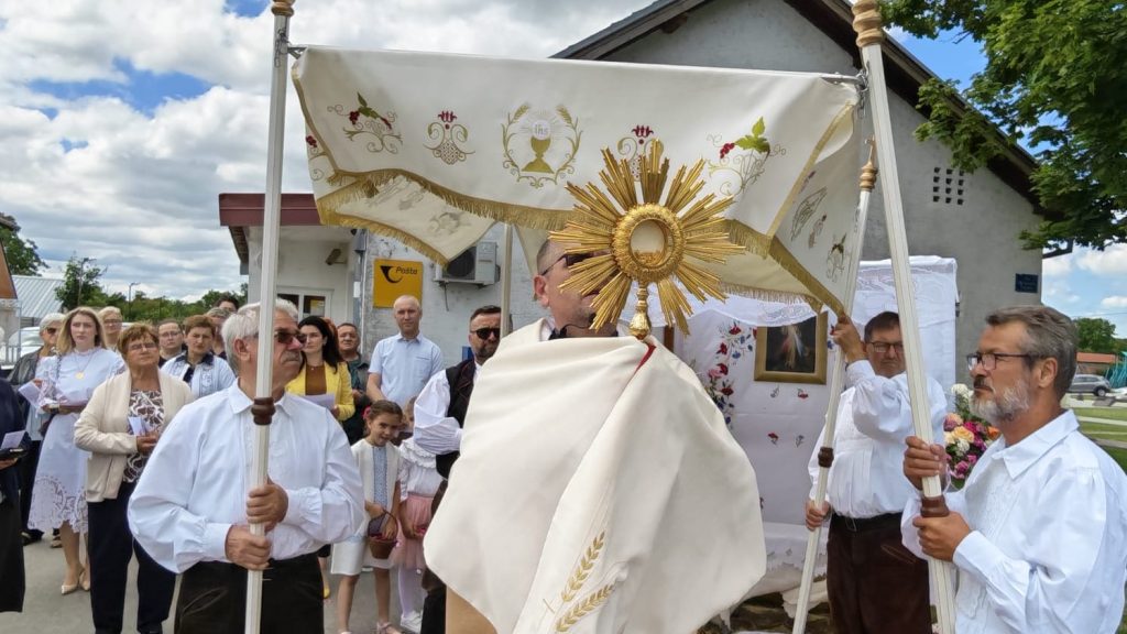 Catholic priest carries a golden monstrance beneath an embroidered ceremonial canopy while a crowd of parishioners in white and pastel attire lines the street in a religious procession.