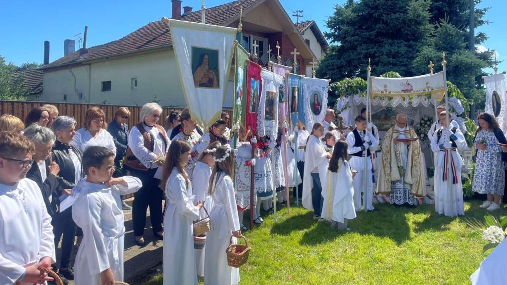 Outdoor religious procession with children in white robes carrying banners and baskets, as a priest leads under ornate canopies/standards.