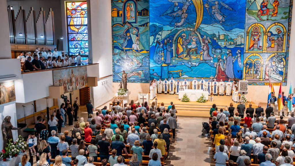 Wide view of a church interior during service: altar with priests, large blue mosaic wall, and a full congregation facing forward.