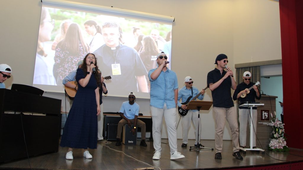 Musicians performing on a stage with a large projected image of a priest behind them as they sing and play instruments.