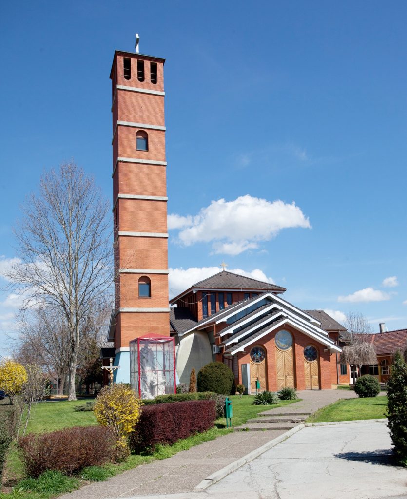 Brick church with a tall, slender red-brick tower beside a red-brick building, under a clear blue sky.