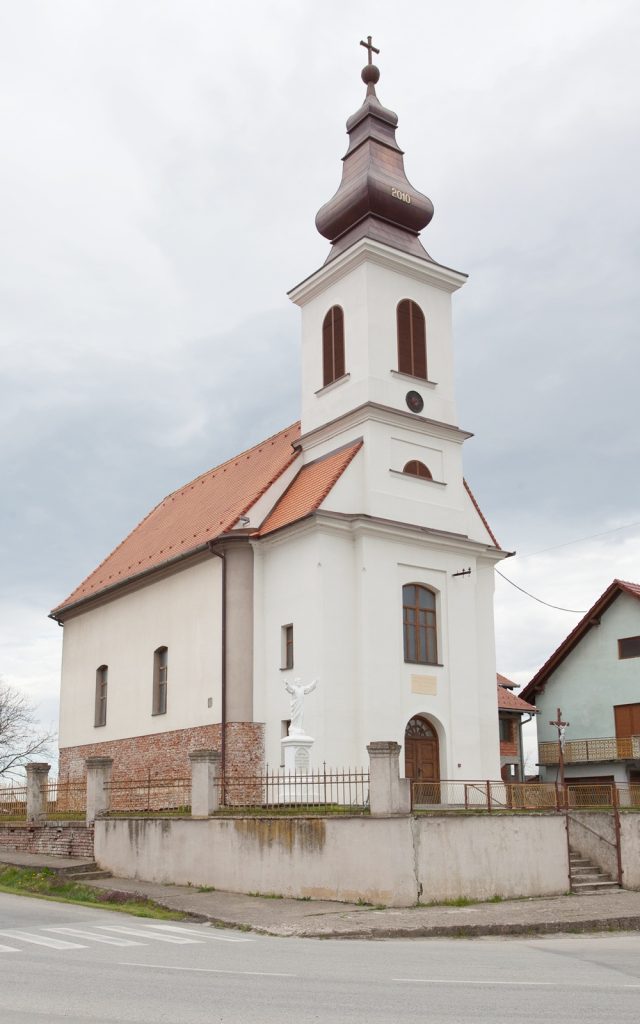 White church with a tall, multi-tiered steeple and orange-tiled roof, statue of an angel near the entrance, fenced yard along the street.