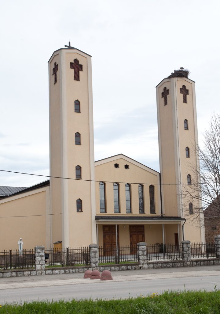 Beige church with two tall towers and crosses, front entrance, fenced yard, and a bird's nest on the right tower top.