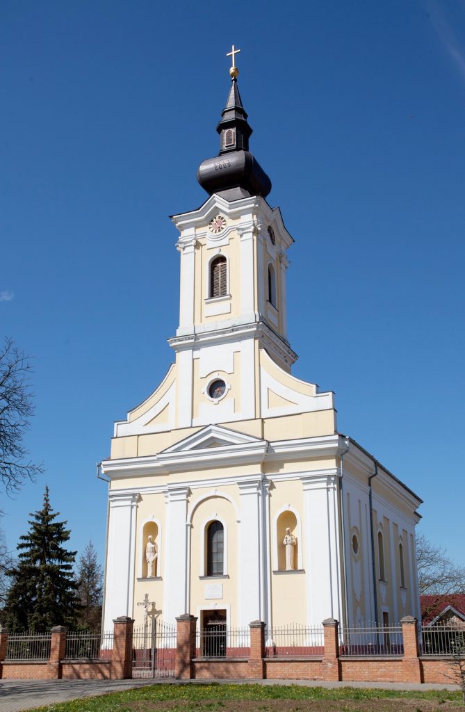 Church with yellow and white façade, arched windows and statues in niches, topped by a tall bell tower and cross against a clear blue sky