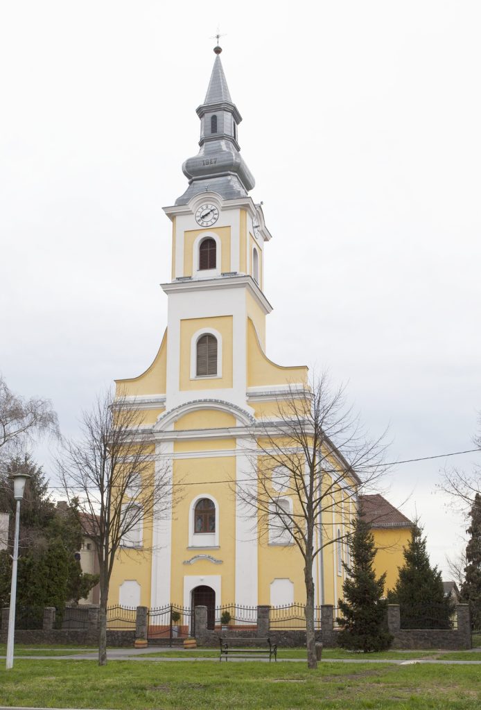 Yellow church with white trim and a tall gray spire housing a clock, set in a park-like area with trees in the foreground.
