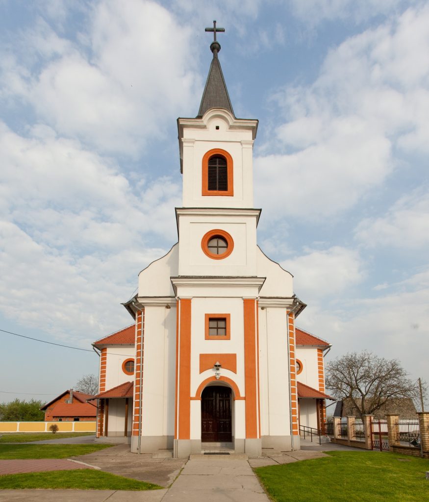 White church with orange trim and a tall steeple topped by a cross against a blue sky and clouds