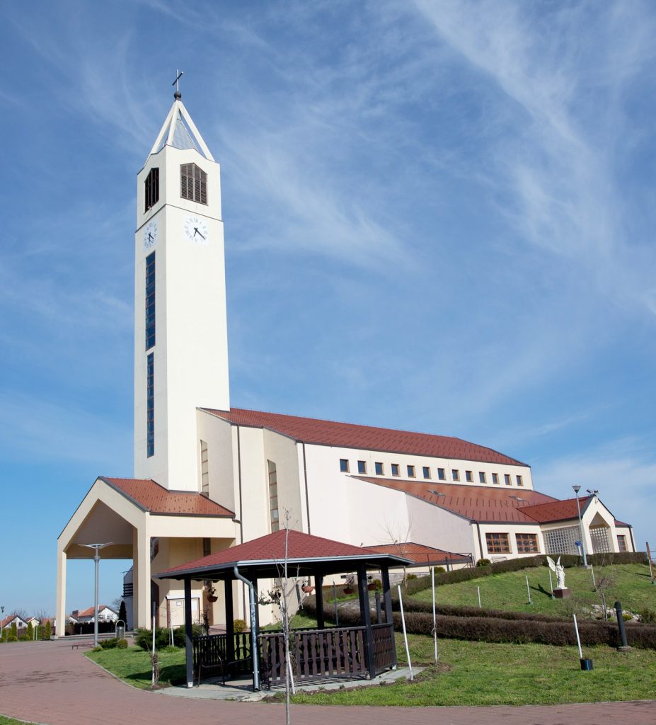 White church with a tall clock-tower reaching into a clear blue sky, red-tiled roofs, and manicured grounds below.