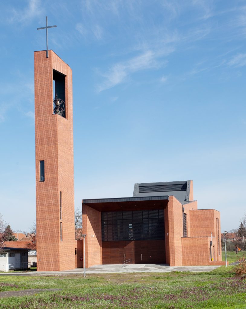 Brick church with a tall rectangular bell tower and cross on top against a blue sky