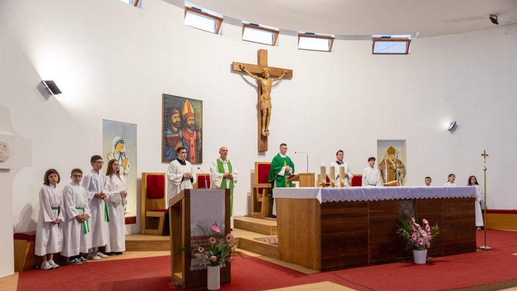 Inside a church sanctuary, priests in green vestments stand at the altar with several altar servers in white robes.