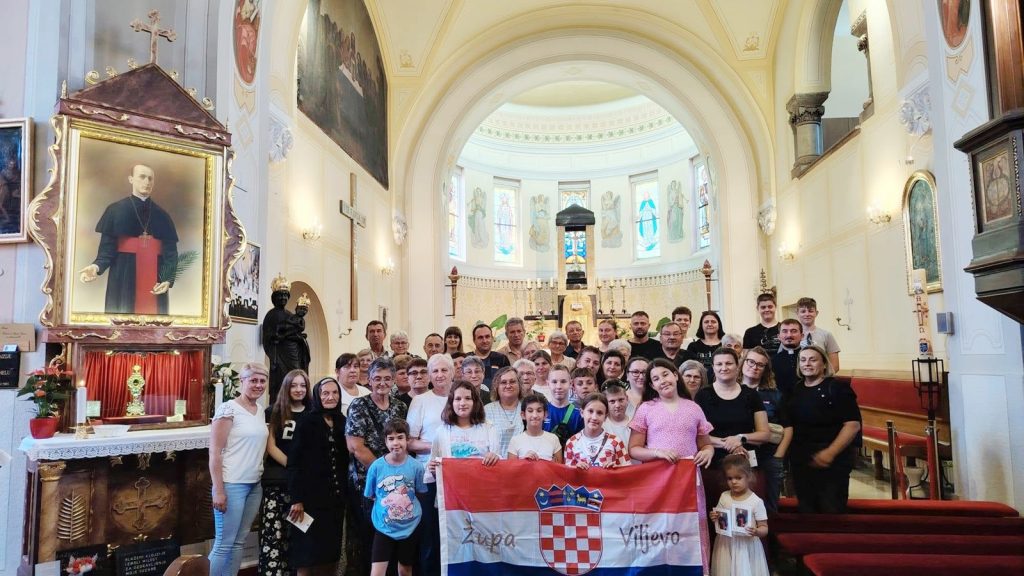 Large group of people posing for a photo inside an ornate church, holding a Croatian flag banner with a coat of arms, stained glass windows in the background.