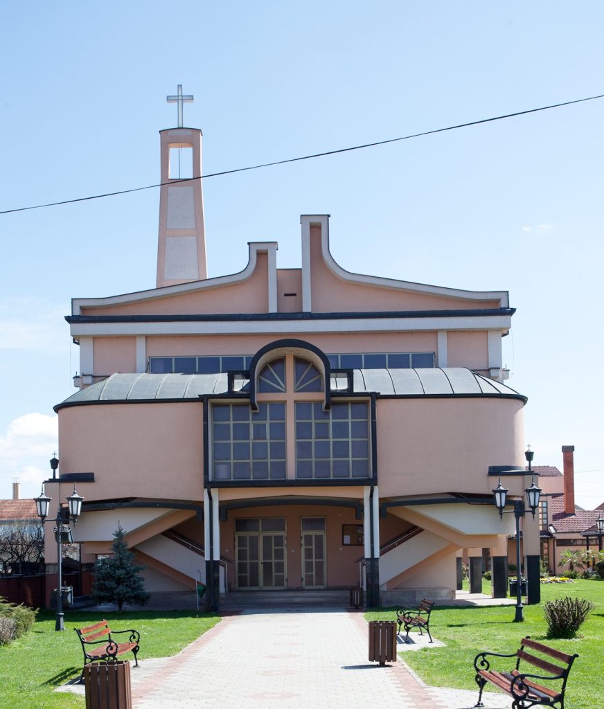 Front view of a peach-colored church-like building with a cross-topped tower and decorative curved roof, entrance in center, benches in foreground