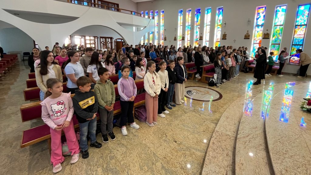 Group of children standing in pews inside a church during a service, with colorful stained-glass windows in the background.