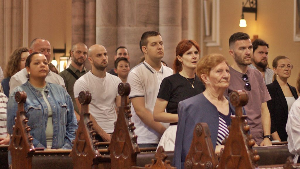 Diverse group of adults standing behind wooden church pews, listening attentively during a service.