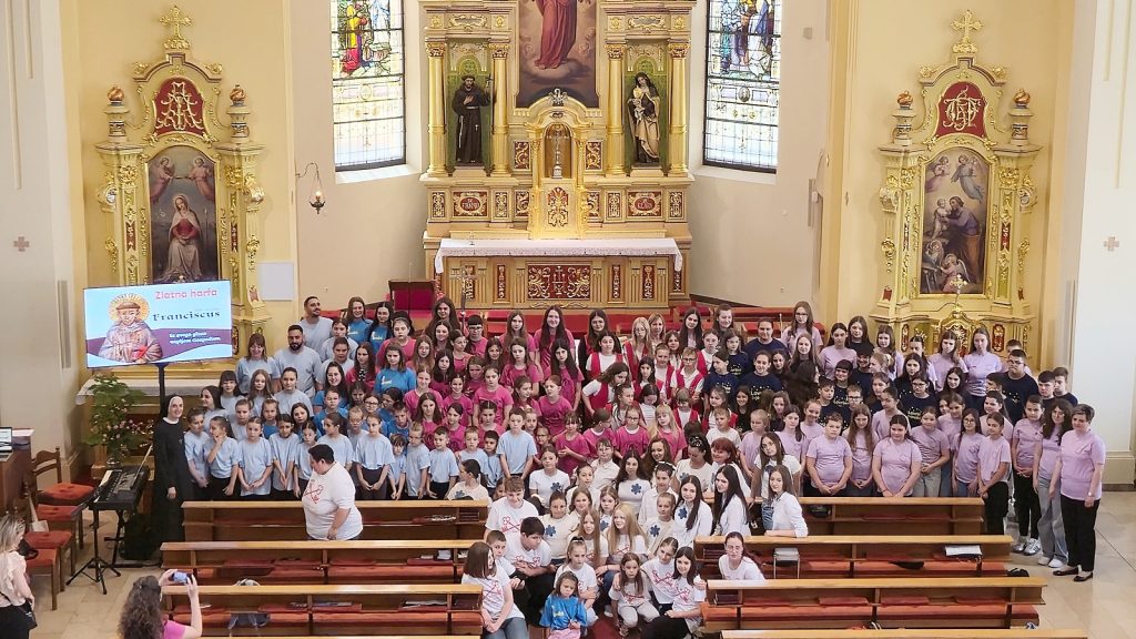 Inside a gilded church sanctuary, a large choir of children and teens in pastel shirts pose for a group photo in front of an ornate altar with statues and stained glass windows.