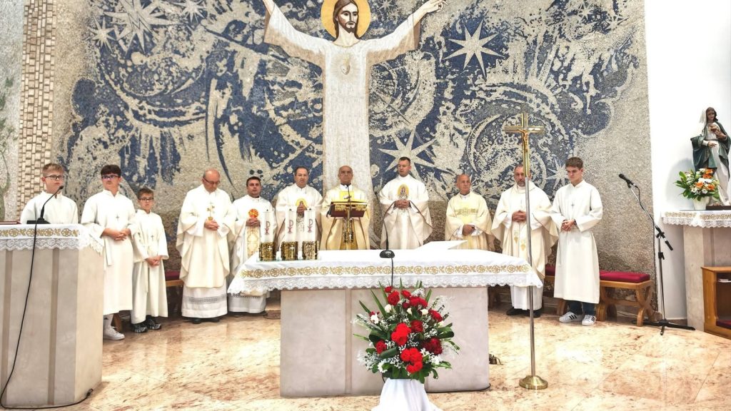 Catholic Mass at the altar with several priests and altar servers in white vestments, a floral arrangement in front, and a large mosaic of Jesus behind.