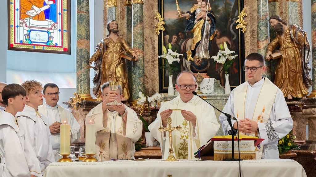 Priests and altar servers in white vestments celebrate Mass at an ornate church altar; one holds up a chalice while another presents the host.
