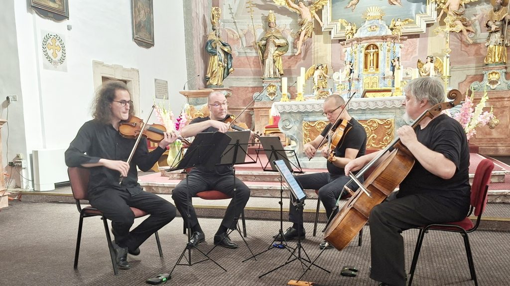 Four musicians in black perform a string quartet inside a ornate church, playing violins and cello with sheet music stands in front of them.