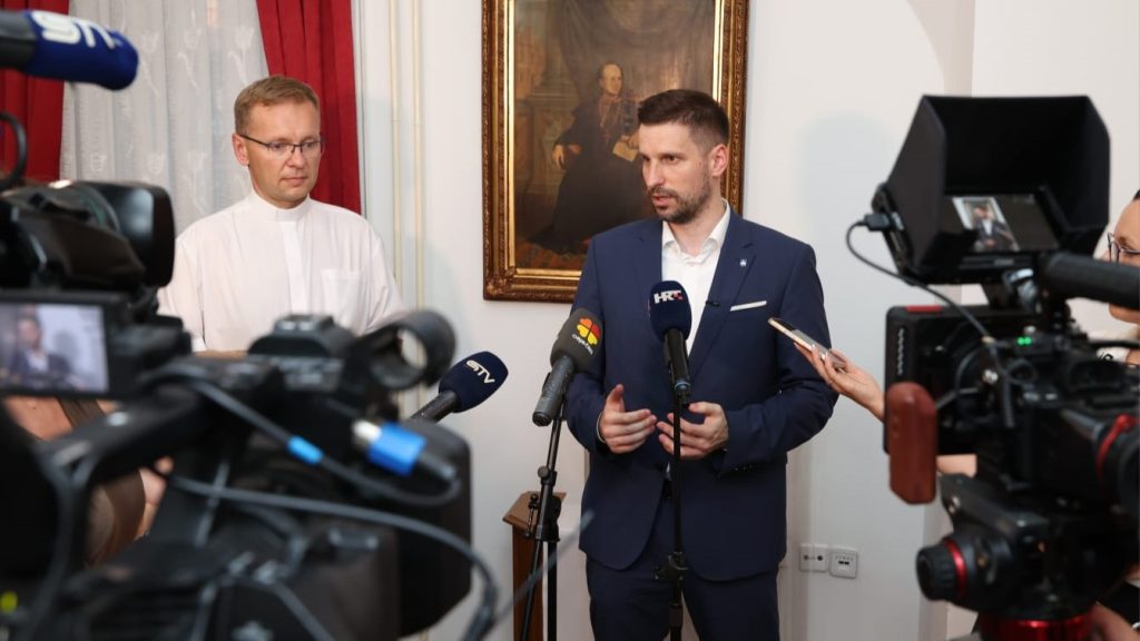 Man in a navy suit giving a press statement as reporters and cameras surround him in a room with a painting behind
