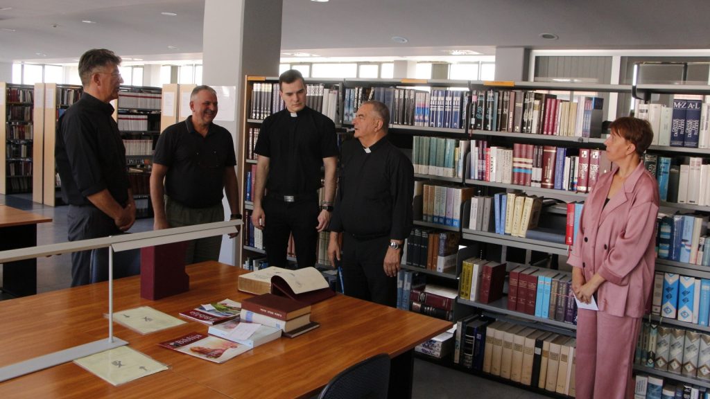 Group of four men, including two priests, and a woman in a pink suit, standing around a library table during a discussion.