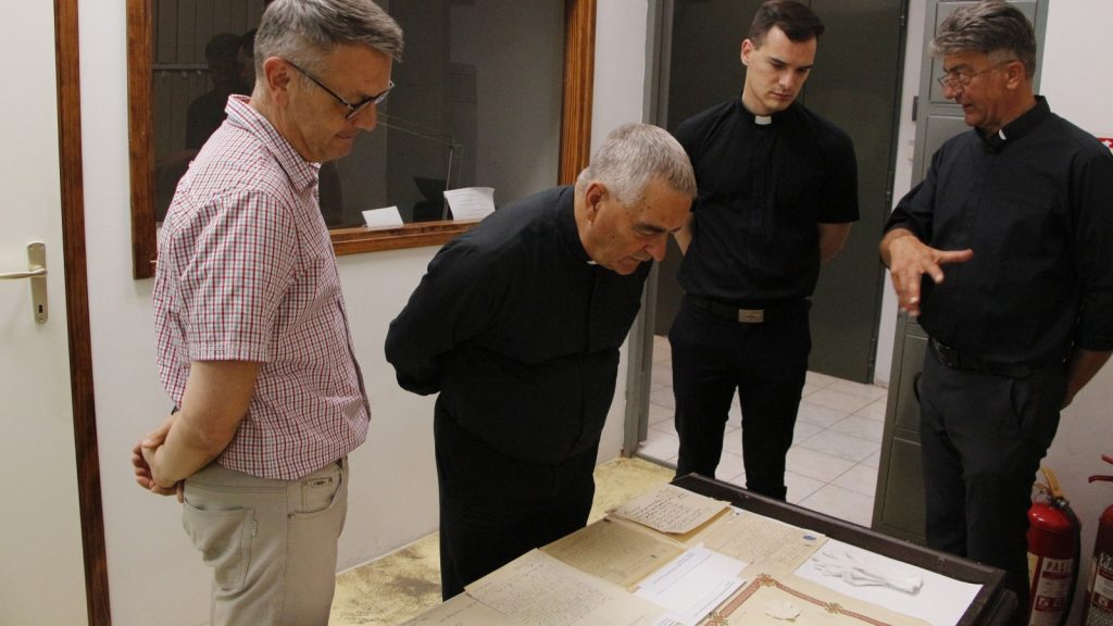 Four men, including two clergy in black shirts, lean over a glass display case examining handwritten documents on a table.