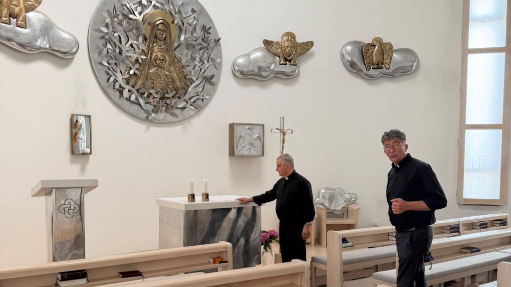 Two Catholic priests in black clerical shirts stand in a bright, minimalist chapel with a marble altar and modern metal reliquaries on the wall behind them.