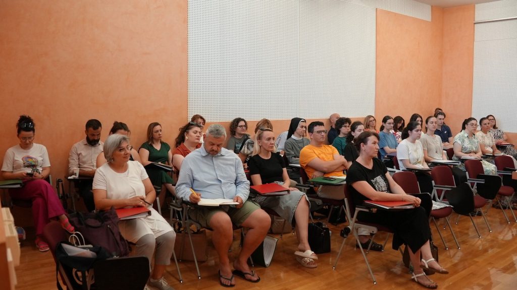 Audience of adults seated in a lecture room, some taking notes, with orange walls and white perforated panels behind them.