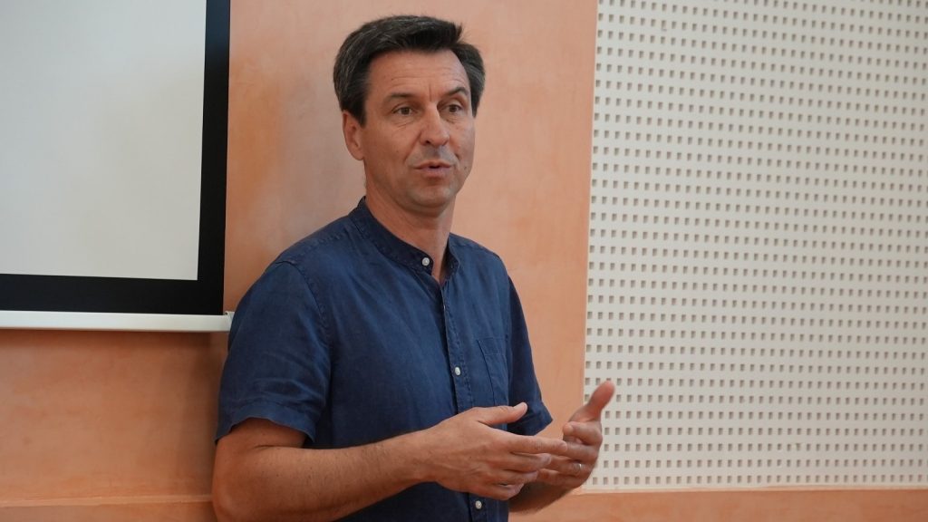Middle-aged man in a blue shirt presenting indoors, using hand gestures while talking.