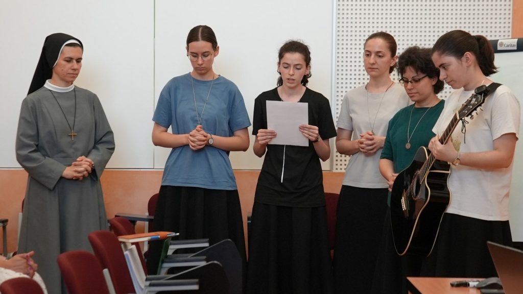 Six young women and a nun stand in a row; one reads from a sheet while another plays an acoustic guitar in a classroom setting.