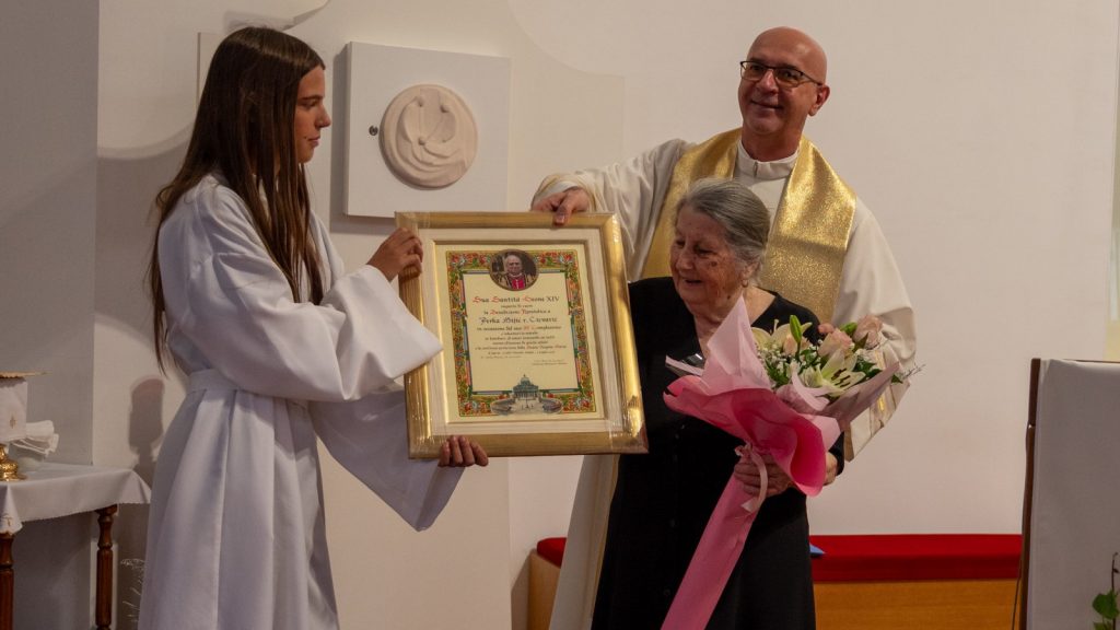 Young girl in a white robe hands a framed certificate to an elderly woman during a religious ceremony, with a smiling priest nearby holding a bouquet crown ceremony.