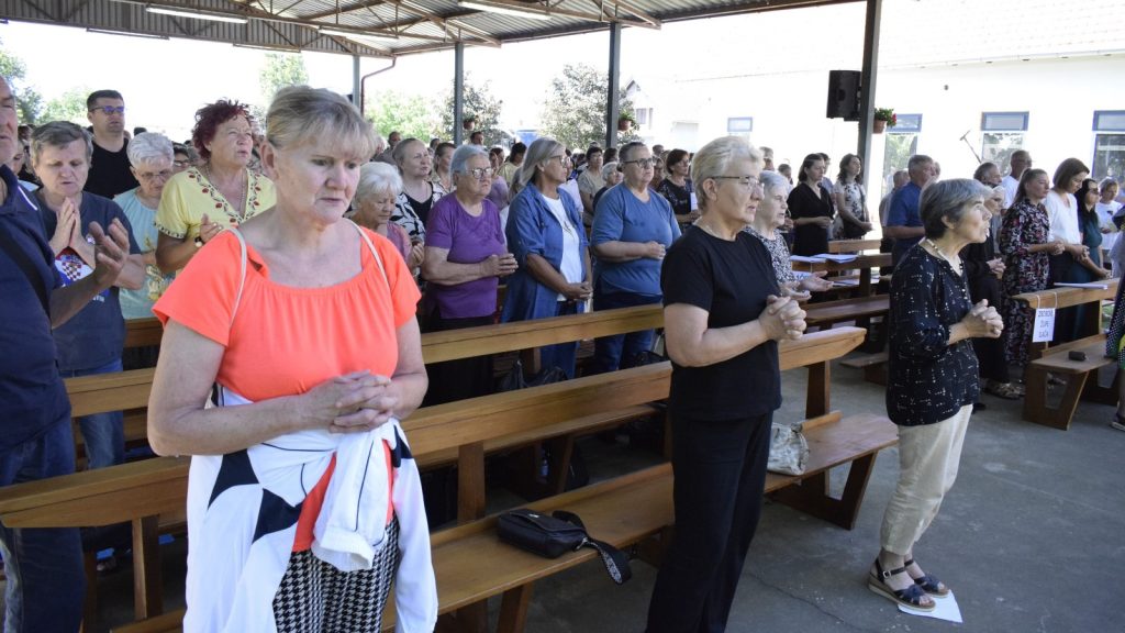 Crowd of mostly seniors standing with hands clasped in a covered outdoor area, benches visible, participants in casual clothing during a gathering.