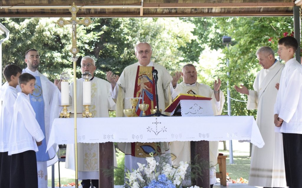 Catholic outdoor Mass with priests at an altar, central bishop blessing with raised hands and microphone nearby.