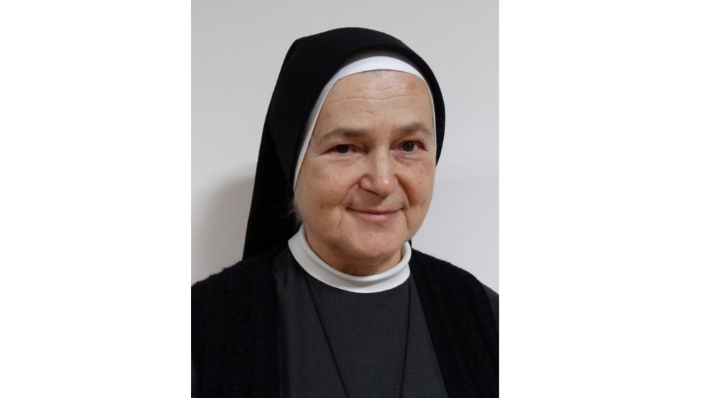 Smiling nun in a black habit and white wimple, facing the camera against a light background