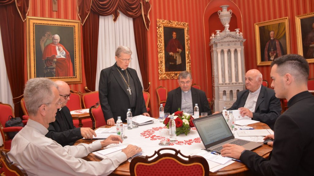 Clergy and laypeople sit around a ornate round table in a red-walled room, discussing documents with a laptop open nearby.