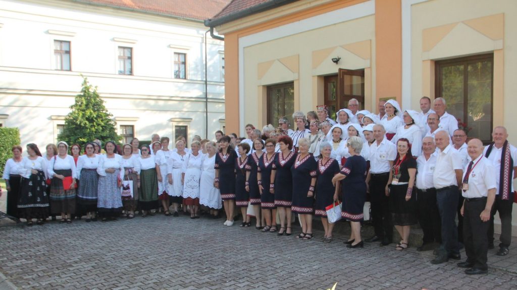 Large group of men and women in traditional folk costumes outside a pale yellow building, posing for a group photo.