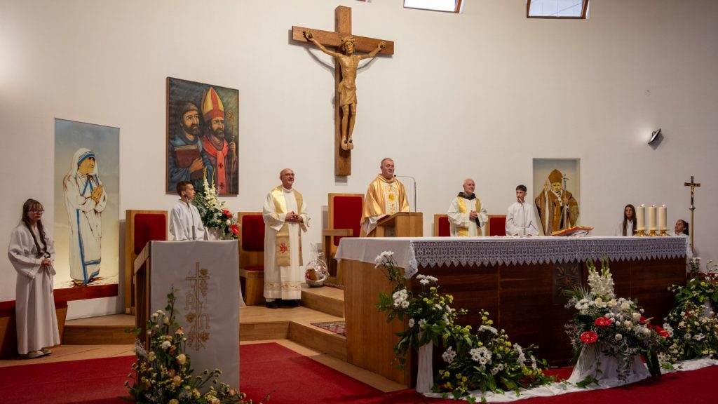 Catholic church altar during Mass with priests at the lectern, crucifix overhead, and floral arrangements on the altar table