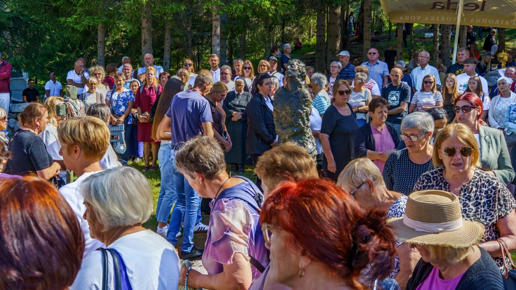 Crowd of people gathered in a park around a statue on a sunny day at an outdoor event—varied ages and outfits visible.