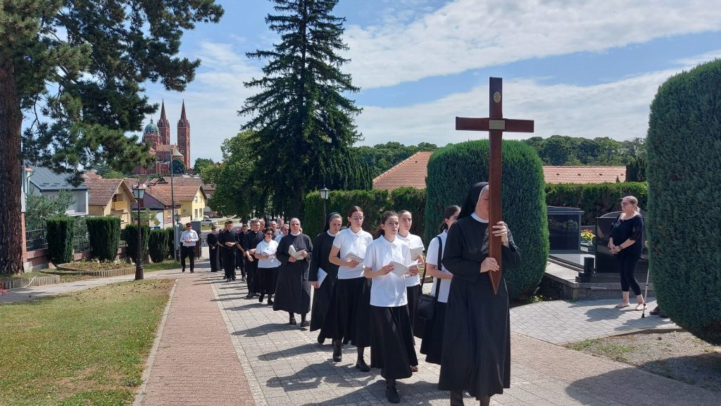 Religious procession of nuns and laypeople walking along a paved path, led by a nun carrying a large wooden cross, with a church and houses in the background under a blue sky.