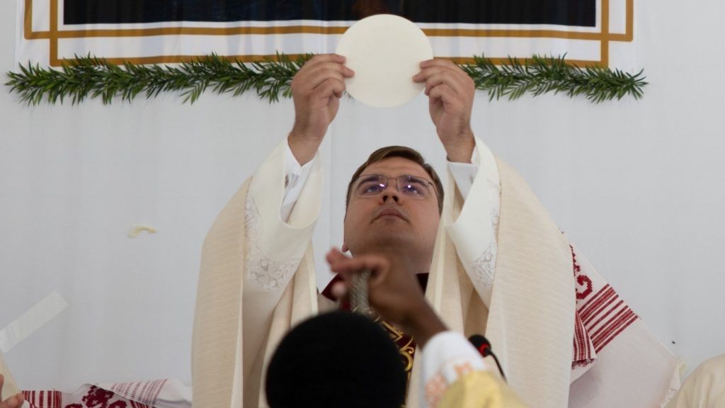 Priest in cream vestments elevates the round communion host during Mass